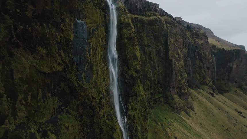 The video is a sweeping shot of a narrow tall waterfall flowing down a sheer mosscovered cliff face the surrounding landscape is rugged and covered in vivid green and brown grasses suggesting a remote wet and mountainous location