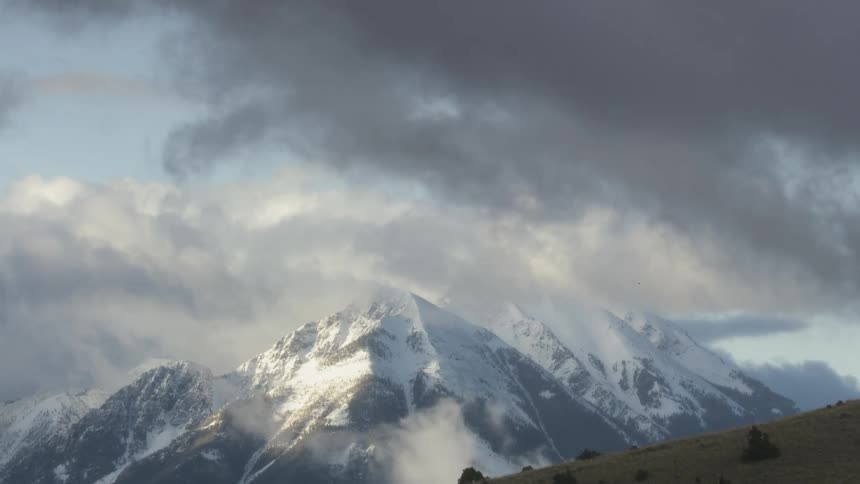 The video is a sweeping shot of a majestic snowcapped mountain range shrouded in clouds dark storm clouds loom over the peaks while patches of sunlight break through to illuminate the white snow