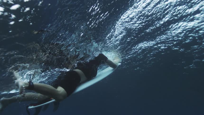 An underwater shot shows a surfer in a dark wetsuit lying prone and paddling a white surfboard across the surface of the ocean water creating bubbles and turbulence above the dark blue depths