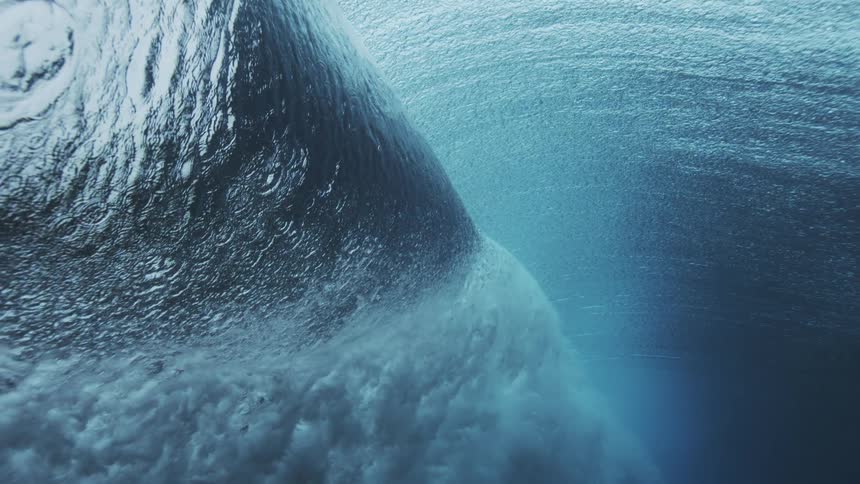 Send an underwater view captures the powerful breaking motion of a wave as it surges across the frame with thick white foam and bright blue light filtering down from the surface