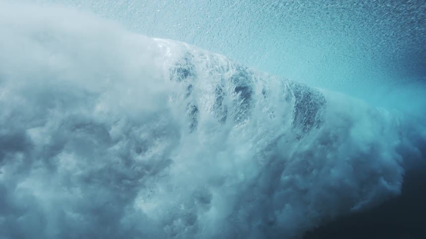 An underwater view shows a dark sloping wall of water with faint ripples then a a billowing cloud of white foam spreads beneath a bright blue surface