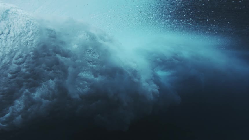 An underwater view captures the foamy turbulent energy of a large ocean wave cresting and breaking as bright light shines through the surface into the deep blue water
