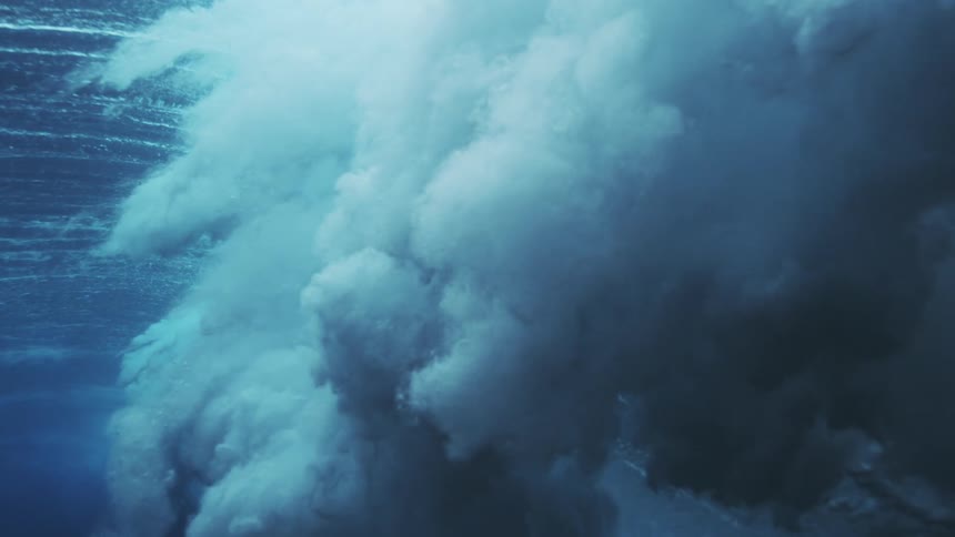 An underwater view shows the foamy underside of a breaking wave curling with bright blue light