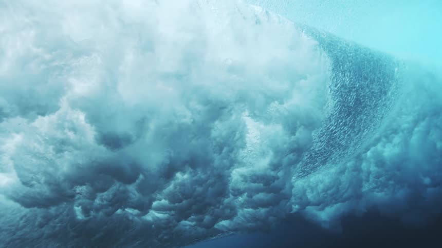 An underwater view captures the powerful churning motion of a giant ocean wave cresting and breaking filling the dark blue water with bright white bubbles and thick foamy turbulence