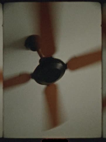 A dark ceiling fan with blurred spinning orange brown blades is mounted on a plain white ceiling in a low angle view