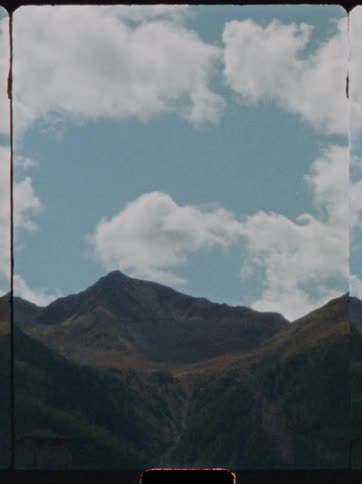 A mountain with a rugged peak draped in patches of grass rises beneath a sky where clouds drift through bursts of sunlight