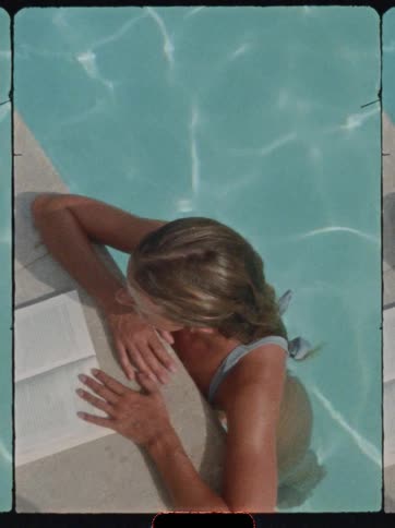 A woman wearing a light blue swimsuit is lying in a swimming pool resting her arms on the white edge while reading an open book as sunlight creates water reflections on her back