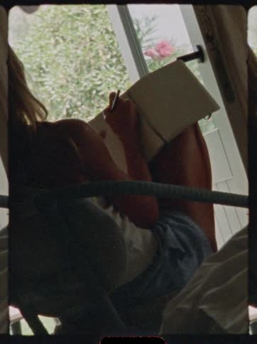 The video is an indoor shot likely captured through a door frame or window showing a person sitting and writing in a notebook the subject is largely silhouetted against the bright green foliage and sunlight visible outdoors