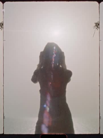 A young woman stands on the beach under the midday sun rubbing her hands through her hair as the light glints off the waves behind her