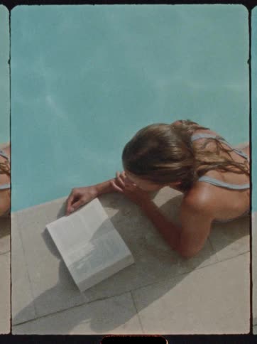 A woman in a pale blue swimsuit is lying on the edge of a swimming pool with a book open next to her resting her head on her arm in the bright sunlight