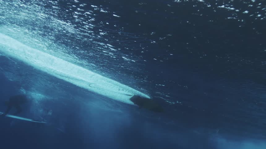 A surfboard glides along the ocean surface seen from beneath as powerful waves crash above churning the water into a mass of bubbles
