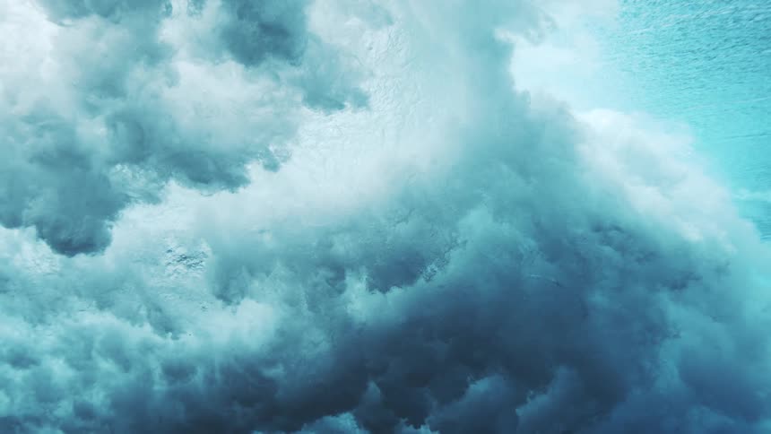 An underwater view shows a large wave cresting and breaking generating white foam and bubbles that expand and rise through the dark blue water