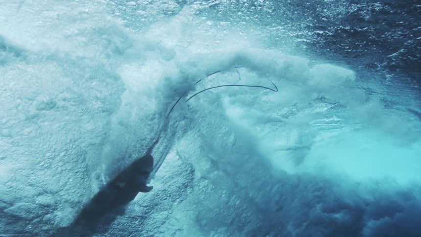 A person rides a surfboard at midday seen from deep beneath the ocean as waves above create foam bubbles and ripple effects across the sunlit surface