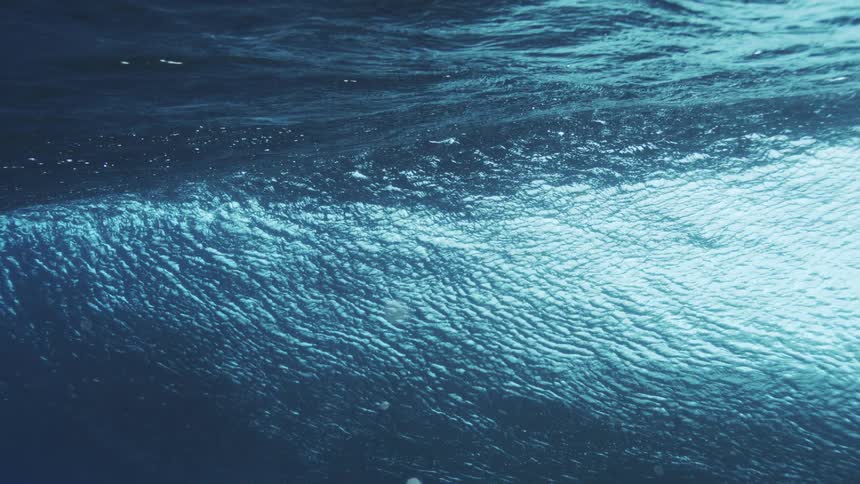 An underwater view shows the foamy underside of a breaking wave curling with bright blue light