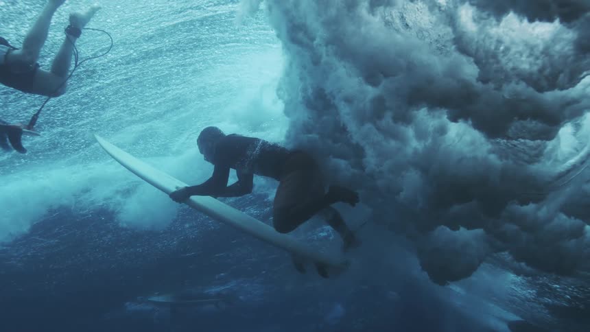 An underwater view captures a surfer duck diving on a white surfboard through a large ocean wave as foam explodes below against the deep blue water