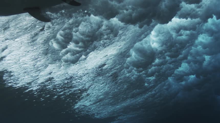 An underwater view captures surfers riding the extreme density and texture of a massive ocean wave breaking with white foam and bubbles engulfing the deep blue frame