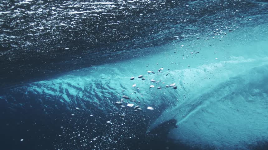 A surfboard glides along the ocean surface seen from beneath as powerful waves crash above churning the water into a mass of bubbles