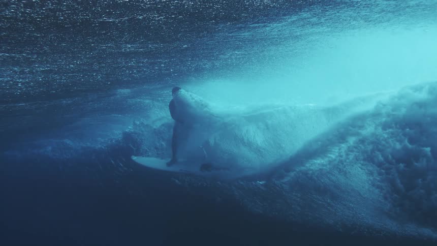 An underwater view shows the interior of an ocean wave as it barrels where a surfer in silhouette rides the side of the water wall on a surfboard as light filters from above