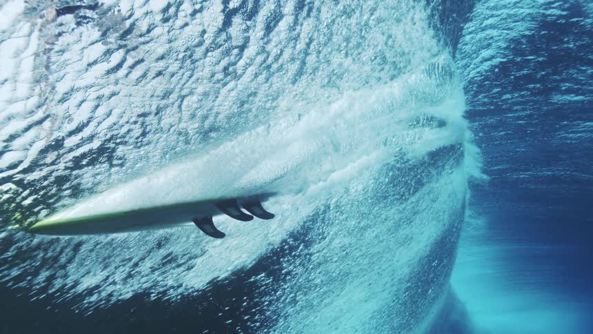 Surfboard cuts through the wave seen from underwater amid bubbles and blue light