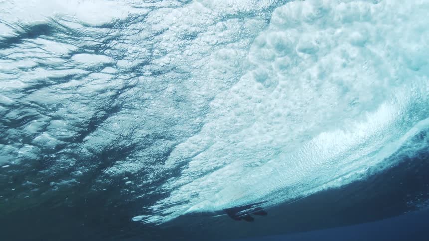 Surfers silhouette glides beneath a wave seen from underwater with bubbles and shifting blue tones