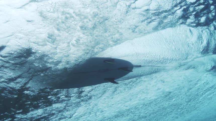 An underwater upwardfacing shot shows a surfboard just beneath the surface of the water leaving a trail of white foam and bubbles behind it while the water surface reflects bright fragmented light