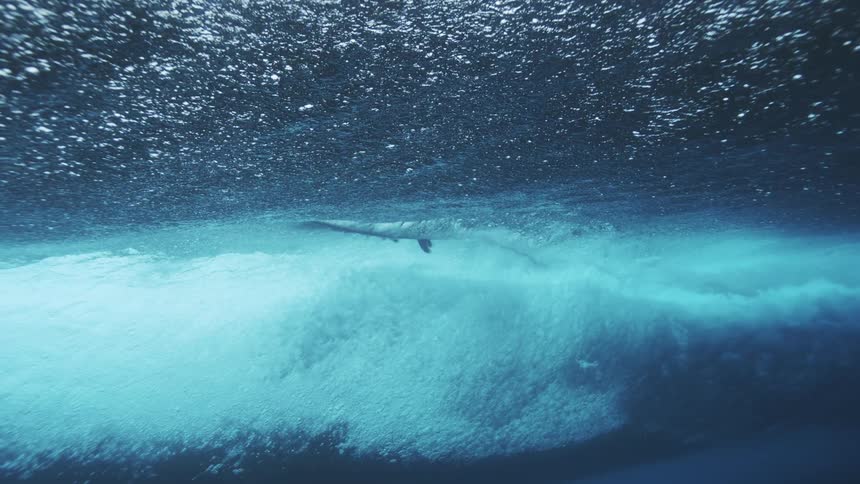 An underwater view shows a surfer riding a wave overhead with the surfboard visible against the bright textured surface of the water and a wall of white bubbles below