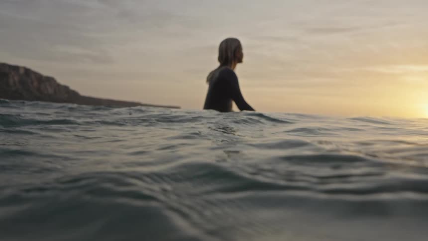 The video shows a lowangle eyelevel shot of a person wading into the ocean as the sun sets the figure is silhouetted against the bright goldenorange horizon with small waves breaking in the foreground