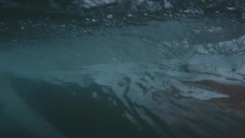 An underwater view captures the powerful flow of a dark blue wave cresting and rushing past the camera with white air bubbles and a dark rock visible on the right