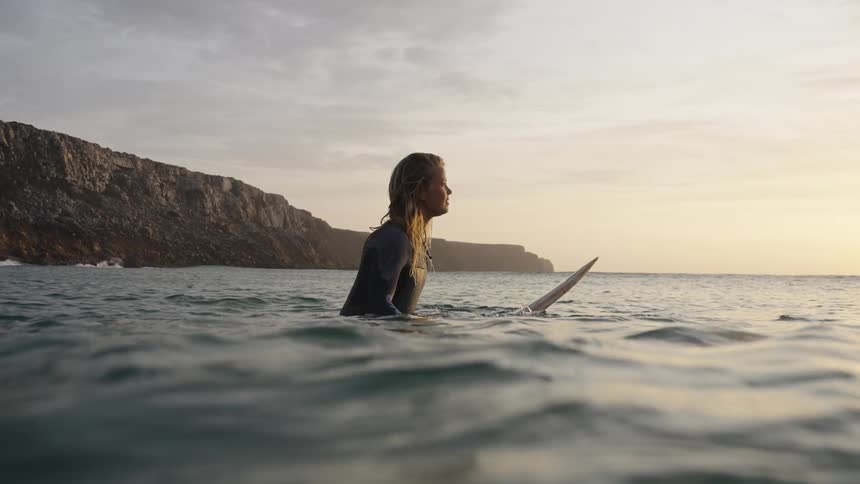 Send a blonde woman wearing a dark wetsuit sits on a surfboard in the ocean turning to smile at the camera as large cliffs form a dark backdrop against the pale sky at sunset