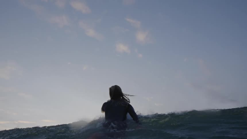 A surfers silhouette is seen from behind paddling in the ocean on a surfboard before quickly duck diving through a large wave at sunset