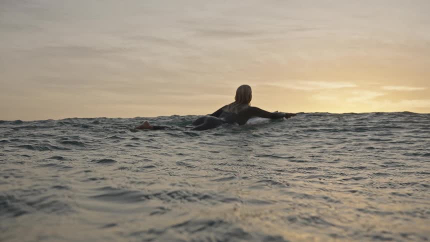 Surfer rests on her board watching the sunset on the golden horizon