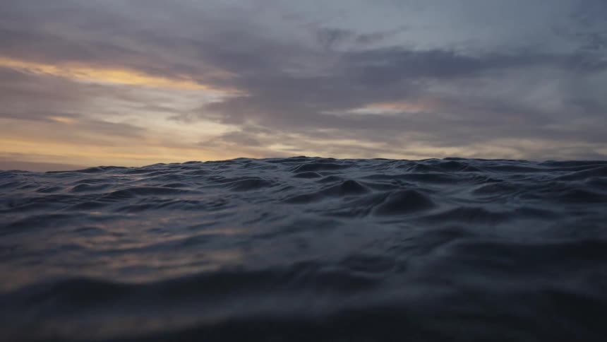 A person is visible as a small dark silhouette standing in the choppy ocean water near the horizon framed by the dark waves and a cloudy sunset sky
