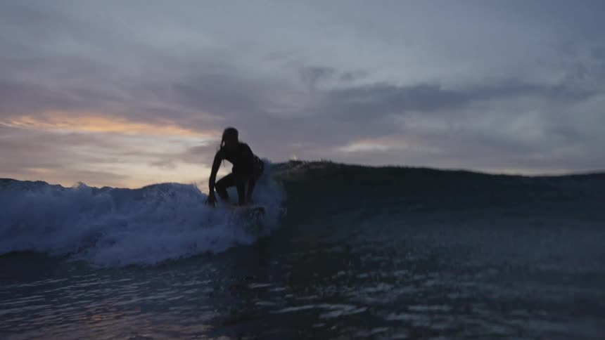 A surfers silhouette is seen riding a small wave on a surfboard crouched low over the water against the bright pale sunset sky