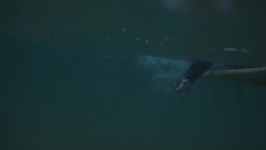 The video is an underwater shot of a surfer wearing a dark wetsuit paddling a surfboard through the deep greenblue water the shot captures the bubbles and disturbance around the boards nose as they glide past the camera