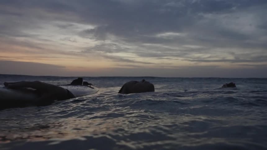 A woman floating on a surfboard silhouetted against the dark water the sky above the horizon is filled with dark clouds and an orange glow