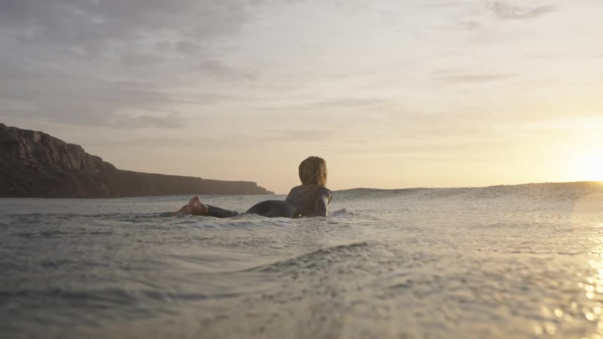 Surfer lying on her board watches the sunset over calm waters near the cliffs