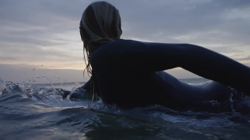 A blonde woman wearing a dark wetsuit lies on a surfboard paddling through choppy dark water toward a coastline of high dark cliffs at sunset