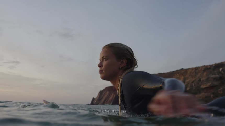 A female surfer paddles on her surfboard another surfer is nearby while gentle swells roll by and cliffs rise in the distance