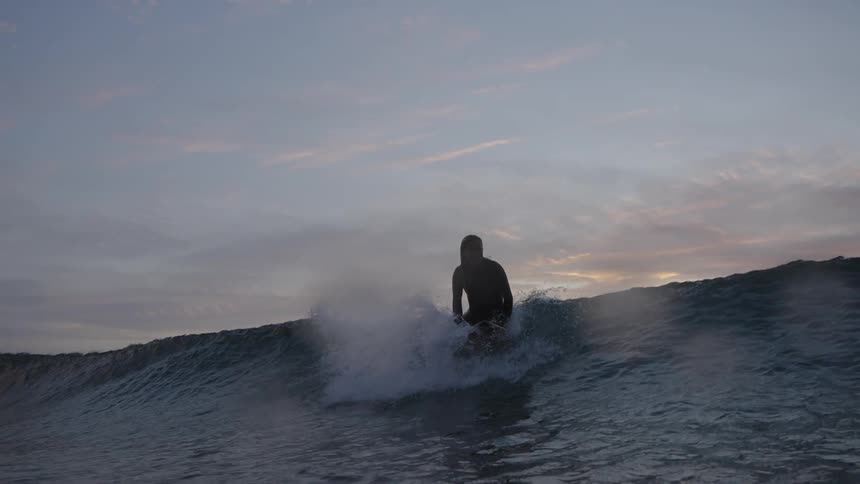 A woman wearing a dark wetsuit stands up to surf a wave into the foreground while another surfer sits nearby all silhouetted against a dramatic sunset sky with dark clouds