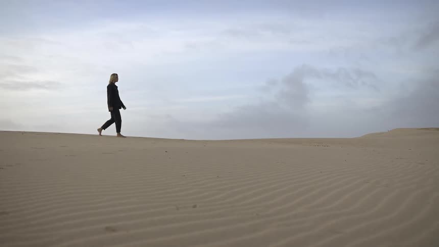 Woman walks alone across sand dunes under a cloudy sky