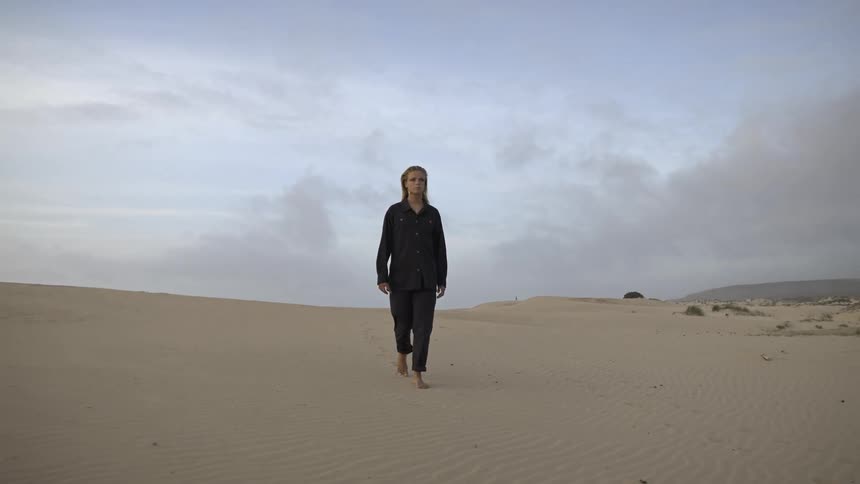 A young woman with blonde hair walks barefoot across desert sand on a cloudy day dressed in soft pajamas as muted light filters through the overcast sky