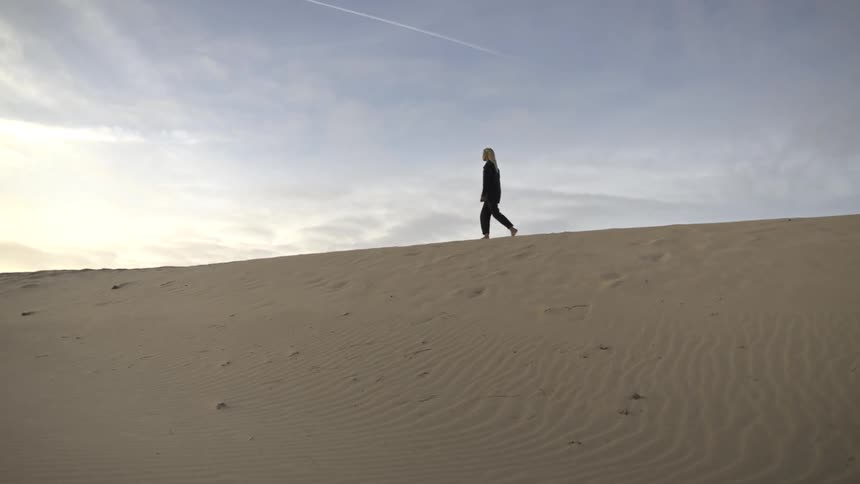A young woman wearing pajamas walks across desert sand at midday