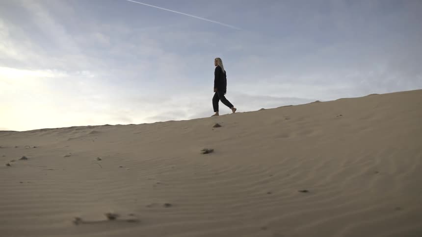 The video shows a woman walking slowly up a sand dune against a pale sky with a thin contrail visible her silhouette is dark against the bright empty landscape creating a sense of isolation or contemplation
