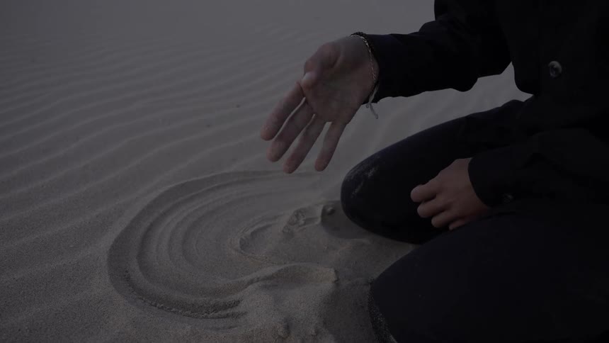 The video is a closeup shot of a persons hand kneeling in the sand the person is wearing dark clothing and is using their hand to draw a circular shape in the fine lightcolored sand