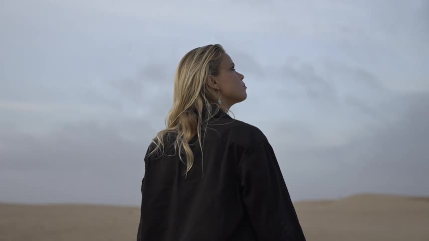 The video shows a woman standing on a beige sandy desert landscape against a pale sky she has blonde hair and is wearing a dark buttondown shirt while looking off to the side with a serious expression