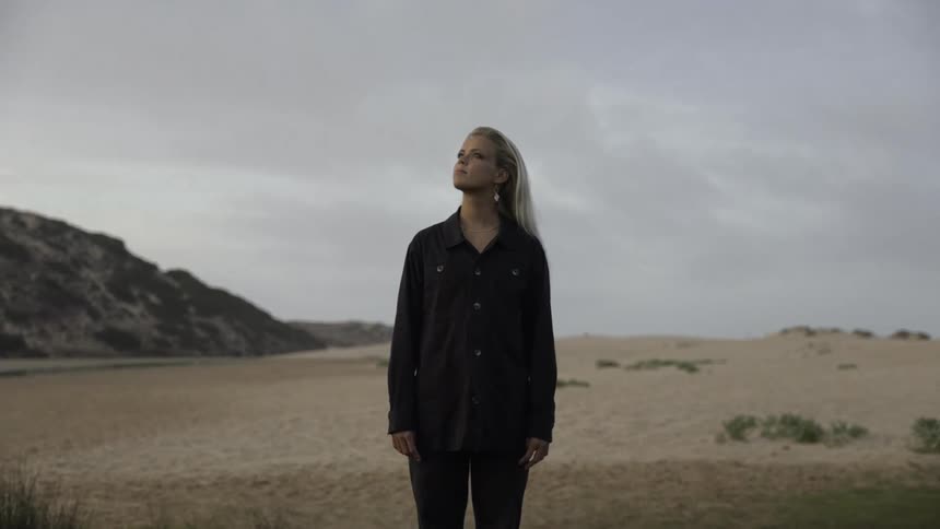 A blonde woman in a dark long sleeved shirt and pants stands barefoot looking up toward the overcast sky in a sandy natural area with dark hills and low green grass