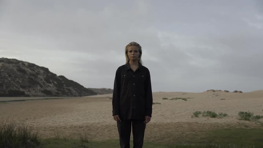 A blonde woman in dark clothing stands barefoot facing the camera in a sandy desert landscape with green bushes dark hills and a pale overcast sky