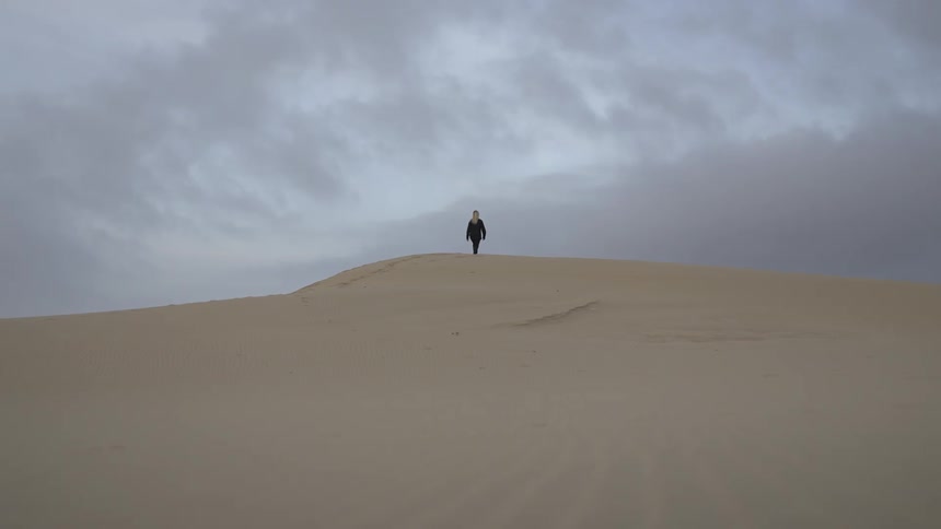 On a cloudy day a young woman walks across desert sand her figure moving quietly beneath the muted sky
