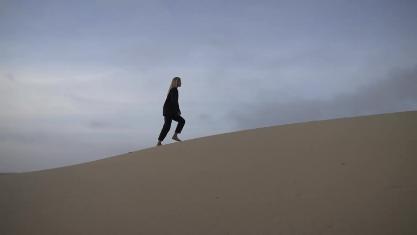 A wide shot shows a woman with long blond hair dressed in dark clothing walking barefoot up the slope of a sand dune against a cloudy pale blue sky