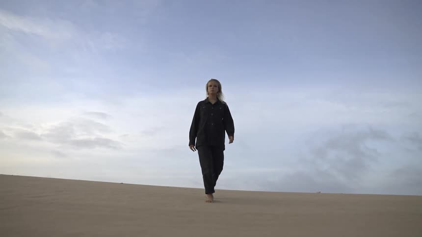 A woman with blonde hair wearing a dark shirt and pants walks barefoot up a large sand dune under a wide pale blue and cloudy sky